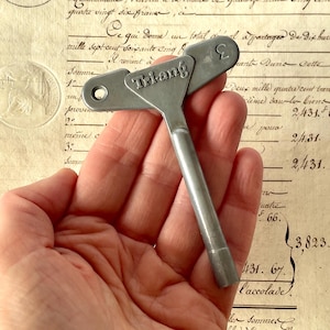 Puede incluir: Una llave de reloj Triang de color plateado, con un mango en forma de T y un agujero circular. La llave tiene la letra "Σ" en la parte superior. La llave se sostiene en una mano, con un documento antiguo en el fondo.