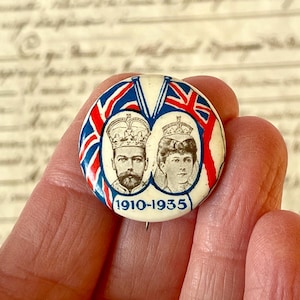 Puede incluir: Un pin redondo vintage con retratos de un rey y una reina, con las fechas 1910-1935. El pin tiene un fondo blanco con diseños de banderas de la Union Jack rojas, blancas y azules. Los retratos están en marcos ovalados.