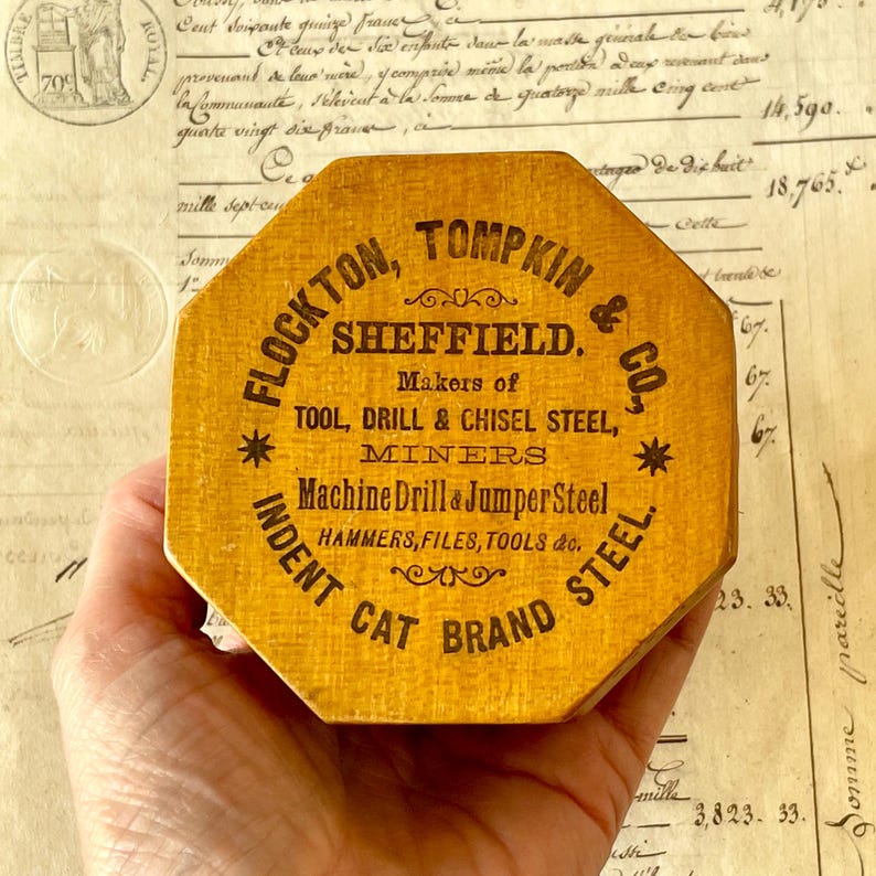 May include: An octagonal wooden box with a yellow finish featuring black printed text that reads FLOCKTON, TOMPKIN & CO. SHEFFIELD. Makers of TOOL, DRILL & CHISEL STEEL, MINERS Machine Drill & Jumper Steel HAMMERS, FILES, TOOLS & CO. INDENT CAT BRAND STEEL.
