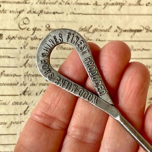 May include: A vintage silver shoe horn with the text "SHOES FINEST PRODUCED WOOD MILNE" embossed on the handle. The shoe horn is held in a hand, with a blurred background of aged paper.