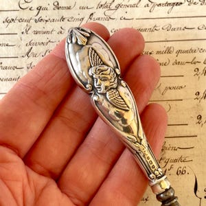 Peut inclure: Gros plan d'une cuillère en argent avec un motif d'ange sur le manche. La cuillère présente des détails complexes et un élément floral décoratif. Le fond est flou et montre du texte manuscrit.