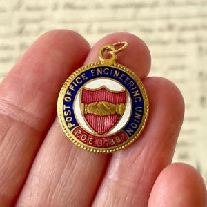 Puede incluir: Un colgante redondo de metal dorado con un borde de esmalte azul con el texto POST OFFICE ENGINEERING UNION y P.O.E.U. 1887. El centro muestra un fondo de esmalte blanco con un escudo rojo que contiene un emblema de un apretón de manos dorado.