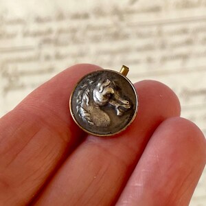 May include: A circular metal button featuring a raised silver-toned relief of a horse head profile centred on a dark background, framed by a thin gold-toned metallic border with a small protruding loop at the top edge for attachment.