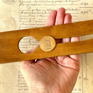 May include: A circular gold-toned metal coin or token featuring an engraved architectural illustration of a cathedral facade with two prominent towers, positioned on a wooden tool with a circular cutout against a vintage document background.