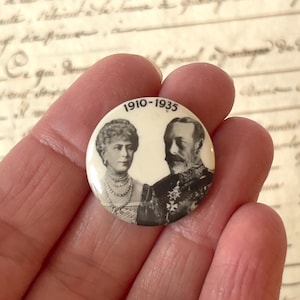 Puede incluir: Un botón redondo vintage con retratos en blanco y negro de un hombre y una mujer, con el texto "1910-1935". La mujer lleva un collar de perlas, el hombre tiene bigote.