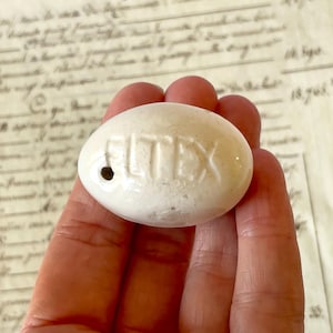 以下が含まれることがあります： 表面に「ELTEX」という文字が浮き彫りにされた、楕円形の白いセラミック製の物体。浮き彫りされた文字の左側には小さな円形の穴があり、表面全体が滑らかで光沢のある仕上がりになっています。