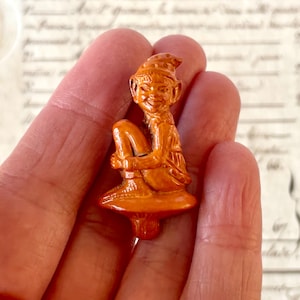Peut inclure: Petite figurine orange représentant un elfe assis avec un chapeau pointu. L'elfe sourit et a les jambes repliées. La figurine est sur une base en forme de champignon. L'elfe se tient les jambes.