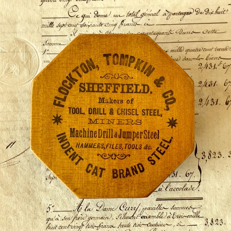 May include: An octagonal mustard-yellow wooden plaque featuring black printed text that reads: FLOCKTON, TOMPKIN & CO. SHEFFIELD. Makers of TOOL, DRILL & CHISEL STEEL, MINERS Machine Drill & Jumper Steel HAMMERS, FILES, TOOLS & CO. CAT BRAND STEEL.