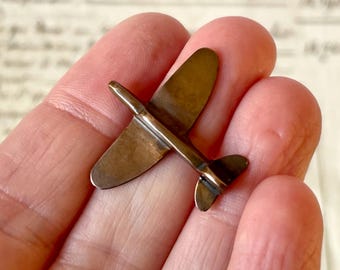 May include: A small, metallic, bronze-coloured aeroplane figurine featuring a single-wing design with a central fuselage and a tail section. The object has a smooth, reflective surface and is shaped like a vintage propeller aircraft.