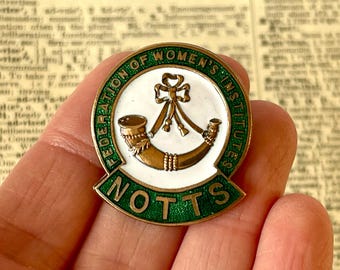 May include: A circular enamel pin with a green border, displaying the text "FEDERATION OF WOMEN'S INSTITUTES" and "NOTTS". The pin's centre has a white background with a gold horn and bow design.