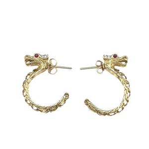 May include: A pair of gold-colored dragon hoop earrings. Each earring features a dragon head with a red gem eye and a curved body. The earrings are set against a white background.