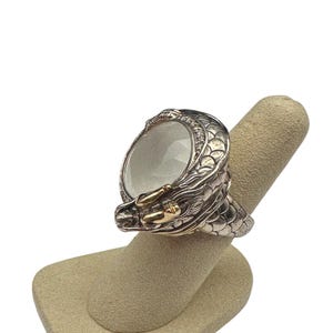 May include: A silver dragon ring with a large, clear, oval-shaped stone. The dragon's head and body are intricately detailed, with gold accents on the face. The ring is displayed on a neutral-colored finger form.