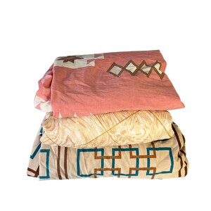 May include: A stack of three folded bed sheets. The top sheet is pink with white and brown geometric shapes. The middle sheet is beige with a leaf pattern. The bottom sheet is beige with brown and teal geometric shapes.