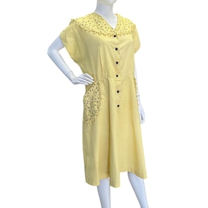 May include: A pale yellow midi dress featuring a wide collar with a scalloped edge and small dark floral print. The dress has short sleeves, a front button placket with dark buttons, and two side pockets with matching scalloped floral trim.