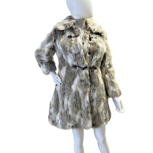 May include: A faux fur coat in shades of gray and white, displayed on a mannequin. The coat features a notched collar, long sleeves, and a flared hem. It has two toggle closures and a mid-thigh length, offering a classic outerwear style.