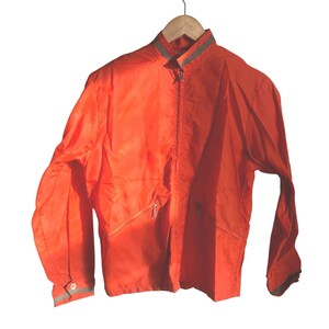 May include: Bright orange jacket with a zipper closure, two front pockets, and a gray reflective stripe on the collar and cuffs. The jacket is hanging on a wooden hanger. The jacket is made of a lightweight material.