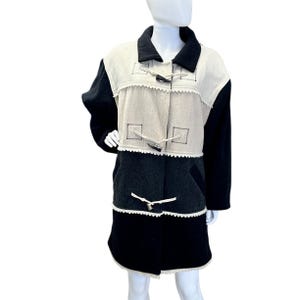 May include: A vintage coat in a color-block design. The coat features black sleeves and a black lower section, with a beige and gray central panel. It has toggle closures and a notched collar. The coat is displayed on a mannequin.