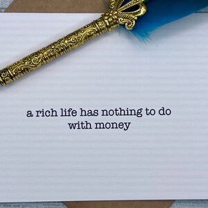 May include: A white card with the quote "a rich life has nothing to do with money" printed in black typewriter font. A gold pen with a blue feather is resting on the card.