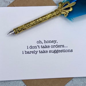 May include: A white card with black text that reads "oh, honey, i don't take orders... i barely take suggestions". A gold pen with a blue feather is on top of the card.