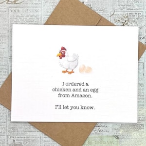May include: A white card with a cartoon illustration of a white chicken laying two eggs. The card has the text "I ordered a chicken and an egg from Amazon. I'll let you know."