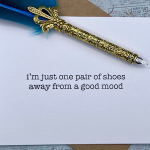 May include: A white card with the text "i'm just one pair of shoes away from a good mood" printed in black. A gold pen with a blue feather is lying on top of the card.