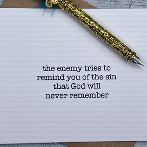 May include: A white card with black text that reads "the enemy tries to remind you of the sin that God will never remember". A gold pen with a blue feather is lying on the card.