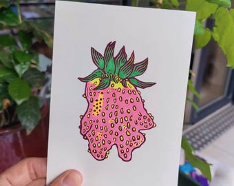Pode incluir: Uma mão segura uma impressão de um morango rosa com a parte superior verde e amarela. O morango tem uma superfície texturizada e manchas amarelas. A obra de arte está em papel branco, com a assinatura do artista e uma data visível.