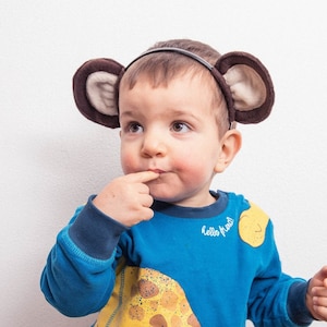 Può includere: Un bambino piccolo che indossa una felpa blu con una stampa di giraffa gialla e il testo "hello friend". Il bambino indossa una fascia marrone con orecchie pelose.