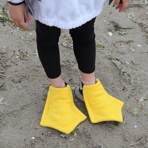 May include: A pair of bright yellow, webbed duck feet shoe covers made of soft fabric. These costume accessories are designed to be worn over footwear, featuring a wide, flat, three-toed shape that mimics the appearance of a duck's webbed feet.