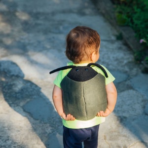 May include: A child wearing a green and black backpack shaped like a beetle. The backpack has large black eyes and a textured surface.