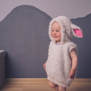 May include: A child wearing a white fluffy lamb costume with pink ears. The costume has a hood and is made of soft, fuzzy material. The child is standing in front of a gray wall.