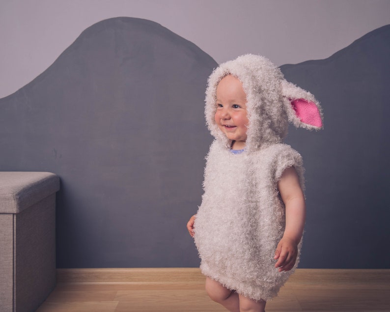 May include: A child wearing a white fluffy lamb costume with pink ears. The costume has a hood and is made of soft, fuzzy material. The child is standing in front of a gray wall.