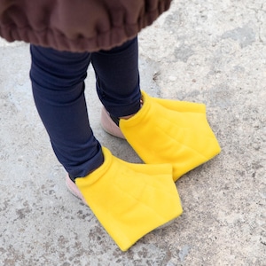 Puede incluir: Un par de fundas de tela tipo forro polar de color amarillo brillante con forma de patas de pato, que se usan sobre el calzado. Las fundas presentan un dise&ntilde;o ancho y palmeado con detalles de costuras que imitan las patas de aves acu&aacute;ticas.