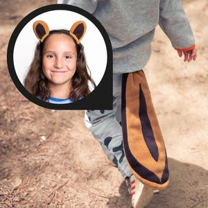 Könnte beinhalten: Ein Kind trägt einen grauen Pullover und eine braune Hose mit einem braunen und schwarzen Eichhörnchen-Schwanz, der an der Rückseite der Hose befestigt ist. Das Kind trägt ein braunes Eichhörnchen-Ohrenband. Das Kind lächelt und schaut in die Kamera.