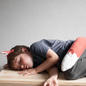 Puede incluir: Un niño con una camisa gris y pantalones grises está durmiendo sobre una superficie de madera. El niño lleva un disfraz de cola de zorro rojo y blanco.