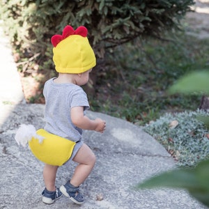 May include: A toddler wearing a yellow and red chicken costume with a white tail feather. The costume includes a hat and a diaper cover.