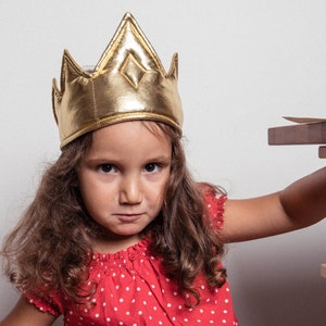 May include: A young girl wearing a gold crown with a serious expression. She is wearing a red and white polka dot dress and has long brown hair.