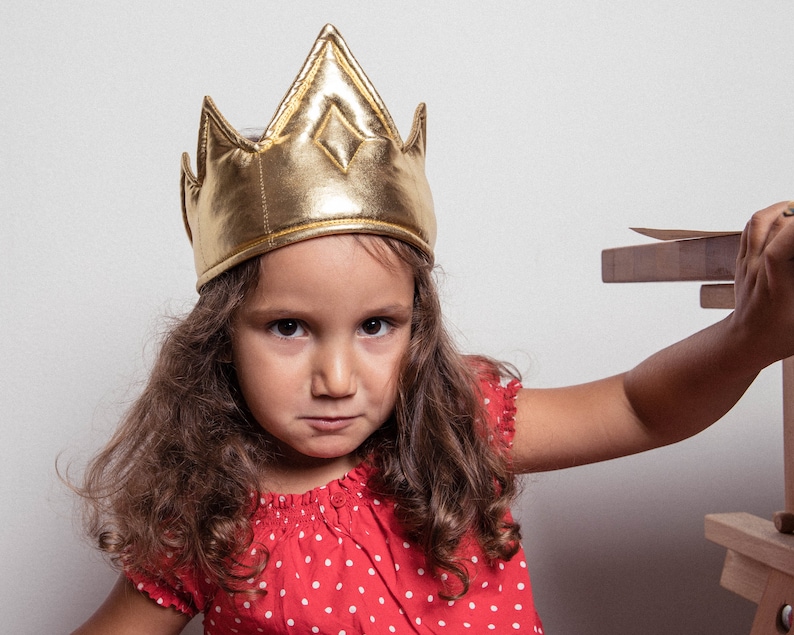 May include: A young girl wearing a gold crown with a serious expression. She is wearing a red and white polka dot dress and has long brown hair.