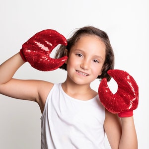 May include: A young person wearing a white tank top is holding up two red sequined lobster claw gloves. The gloves are made of red sequins and have a black elastic band around the wrist.
