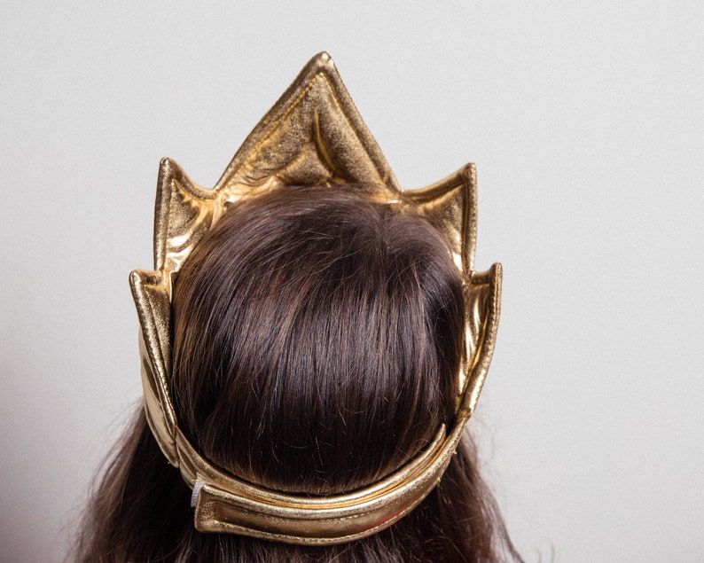 May include: A gold crown headband with a pointed top and a wide band. The headband is made of a soft, flexible material and is designed to fit comfortably on the head.