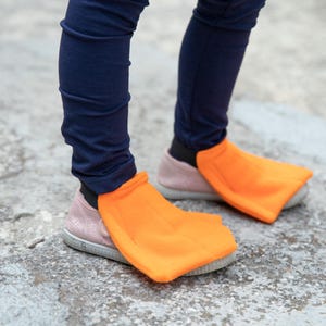 May include: A child's feet wearing orange duck feet shoe covers over pink sneakers and navy blue leggings. The bright orange covers are shaped like duck feet, adding a playful touch to the outfit. The shoes are on a gray concrete surface.