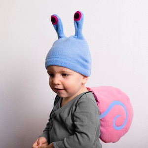 May include: A child wearing a blue fleece snail costume with pink shell and large eyes. The costume is perfect for a Halloween party or a playdate.