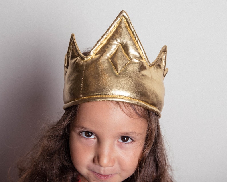 May include: A young person wearing a gold crown with a diamond-shaped design on the top. The crown is made of a soft, padded material.