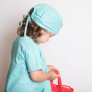 May include: A young child dressed as a doctor in a light blue scrub top and matching hat. The child is holding a red bag.