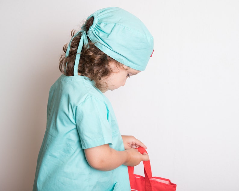 May include: A young child dressed as a doctor in a light blue scrub top and matching hat. The child is holding a red bag.