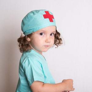 May include: A young child dressed as a doctor, wearing a light blue scrub top and a matching hat with a red cross on it.