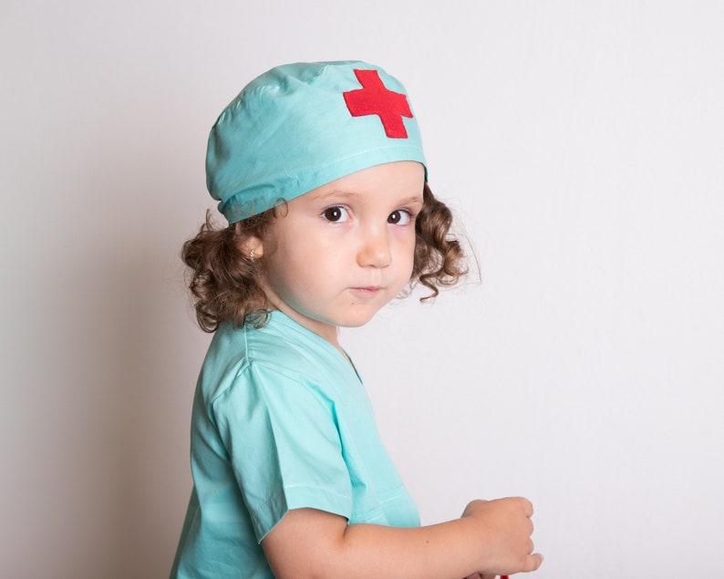 May include: A young child dressed as a doctor, wearing a light blue scrub top and a matching hat with a red cross on it.