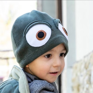 May include: A dark green fleece hat featuring large, cartoon eyes. The eyes are white with brown and black details. The hat is designed to look like a creature's face, suitable for children's costumes or general wear.