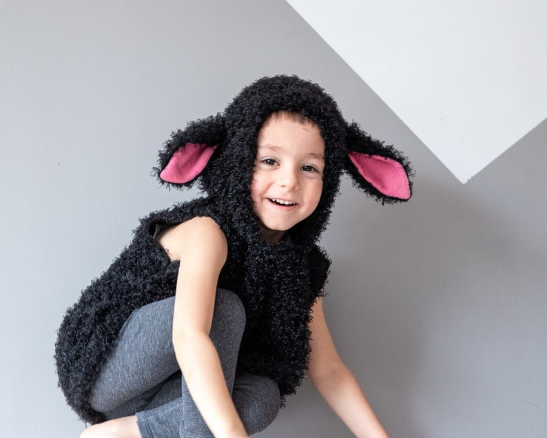 May include: A child wearing a black sheep costume with pink ears. The costume is made of fluffy material and has a hood. The child is smiling and looking at the camera.