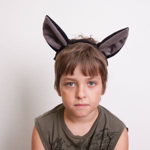 May include: A young person wearing a gray and black donkey ear headband. The headband is made of soft fabric and has a black elastic band that goes around the head.